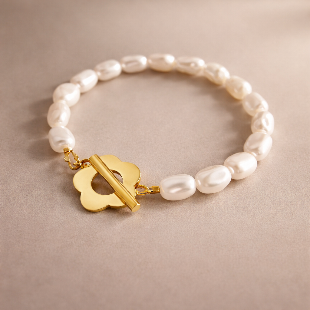 Ein elegantes Armband liegt flach auf einem hellbeigen, glatten Untergrund. Das Armband besteht aus gleichmäßig aneinandergereihten, cremeweißen Süßwasserperlen, die leicht unregelmäßig geformt sind und dadurch natürlich wirken. Am vorderen Teil des Armbands befindet sich ein glänzender, goldfarbener Verschluss in Form einer stilisierten Blüte mit abgerundeten Blütenblättern. Durch die Mitte dieser Blüte verläuft ein glatter, zylindrischer Goldsteg, der als Knebelverschluss dient. Das Gold ist hochglanzpoli