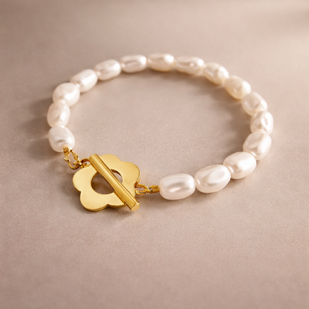 Ein elegantes Armband liegt flach auf einem hellbeigen, glatten Untergrund. Das Armband besteht aus gleichmäßig aneinandergereihten, cremeweißen Süßwasserperlen, die leicht unregelmäßig geformt sind und dadurch natürlich wirken. Am vorderen Teil des Armbands befindet sich ein glänzender, goldfarbener Verschluss in Form einer stilisierten Blüte mit abgerundeten Blütenblättern. Durch die Mitte dieser Blüte verläuft ein glatter, zylindrischer Goldsteg, der als Knebelverschluss dient. Das Gold ist hochglanzpoli